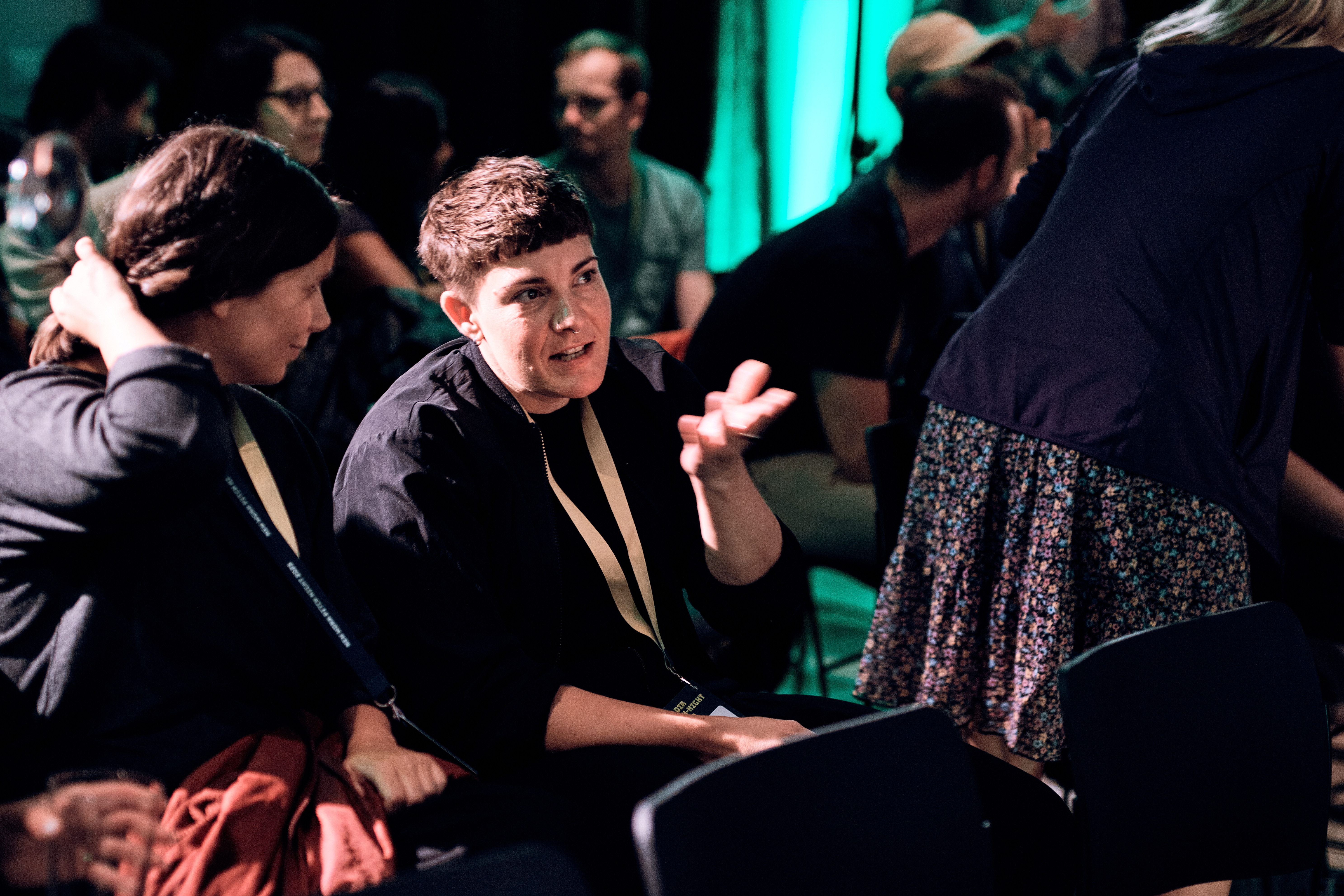 Zwei Personen unterhalten sich im Publikum, während andere im Hintergrund bei schummrigem blau-grünem Licht sitzen oder sich bewegen.